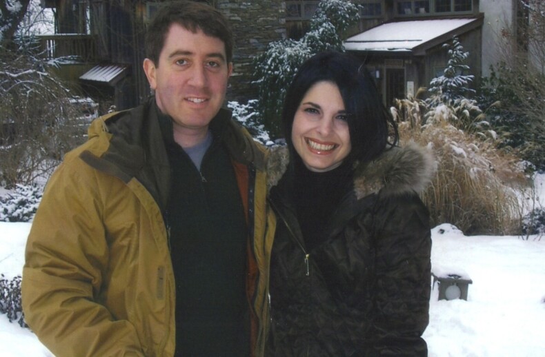 A man and woman stand outside in a snowy garden, smiling, embodying the warmth of social emotional learning. Both wear brown winter coats. Behind them is a house with stone and wood elements, surrounded by snow-covered trees and shrubs.