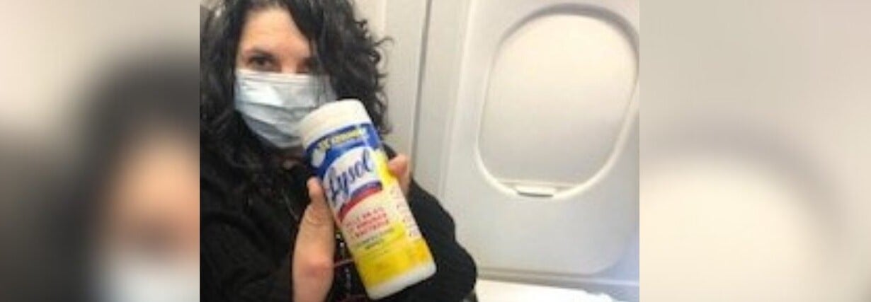 A person wearing a face mask holds a container of disinfecting wipes, sitting by an airplane window, reflecting on the insights gained from their recent SEL curriculum training.