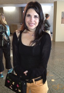 A woman with long dark hair stands indoors, smiling at the camera. She is wearing a black top and cardigan with light-colored pants and holds a black bag with floral designs, perfectly suited for diversity programs. People are visible in the background, suggesting lively interactions.