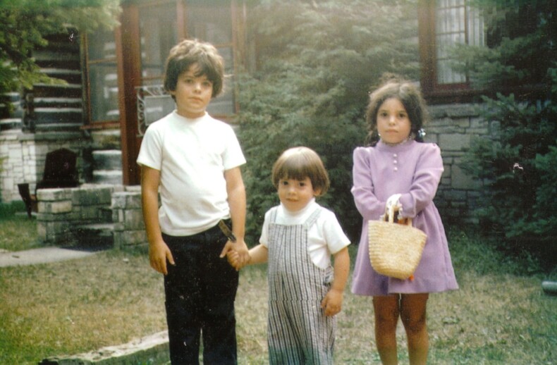 Three children, engaged in social emotional learning activities, stand outdoors in front of a rustic house. One boy in a white shirt holds the hand of a younger child in striped overalls. A girl in a purple dress holds a wicker basket. Trees and garden elements surround the scene.