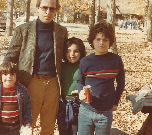 In an autumn park, a man in sunglasses and a blazer stands with three children amid fallen leaves. Embracing the spirit of social-emotional learning activities, one child casually holds a soda can as they enjoy this bonding moment under mostly bare trees.