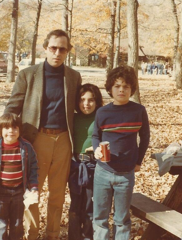 In an autumn park, a man in sunglasses and a blazer stands with three children amid fallen leaves. Embracing the spirit of social-emotional learning activities, one child casually holds a soda can as they enjoy this bonding moment under mostly bare trees.