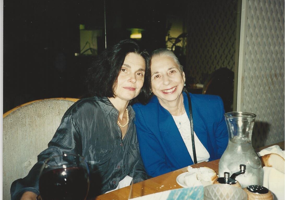 Two women sit closely together at a dining table, smiling at the camera. One wears a dark blouse and pearls, the other a blue blazer. As they enjoy their evening, they discuss engaging social-emotional learning activities over a glass of red wine and water carafe set before them.