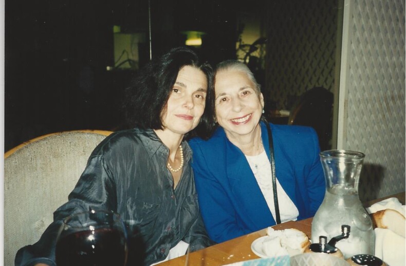 Two women sit closely together at a dining table, smiling at the camera. One wears a dark blouse and pearls, the other a blue blazer. As they enjoy their evening, they discuss engaging social-emotional learning activities over a glass of red wine and water carafe set before them.
