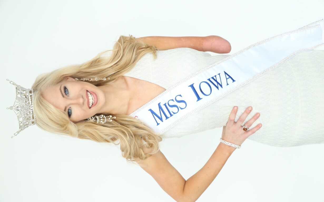 A woman wearing a white dress, a "Miss Iowa" sash, and a tiara smiles at the camera, embodying the spirit of diversity programs. She has long blonde hair, elegant jewelry, and one arm partially missing, promoting inclusivity through her grace and resilience.