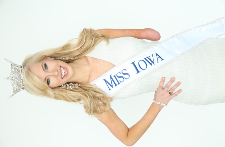 A woman with long blonde hair wearing a tiara and a "Miss Iowa" sash smiles at the camera, embodying the spirit of diversity programs. She is dressed in a light-colored, sleeveless gown and has a prosthetic arm.