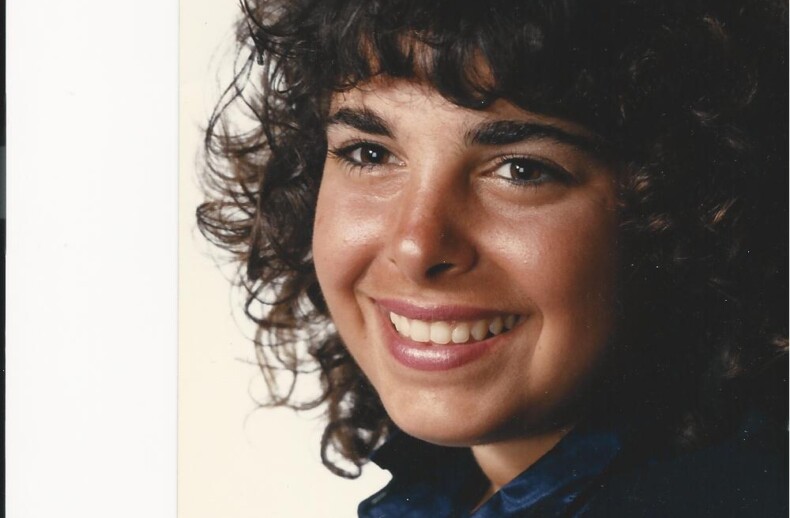 A woman with curly hair, embodying the spirit of diversity programs, smiles warmly at the camera. She is wearing a dark blue blouse against a plain background, radiating confidence and inclusivity.