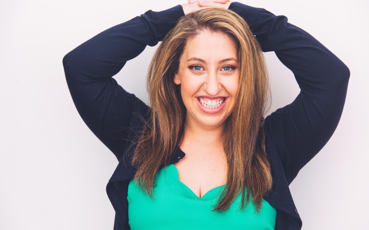 A woman with long brown hair smiles brightly against a white background. She wears a green top and black cardigan, her hands resting on her head. Her demeanor radiates warmth, much like an engaging facilitator of social emotional learning activities or diversity programs.