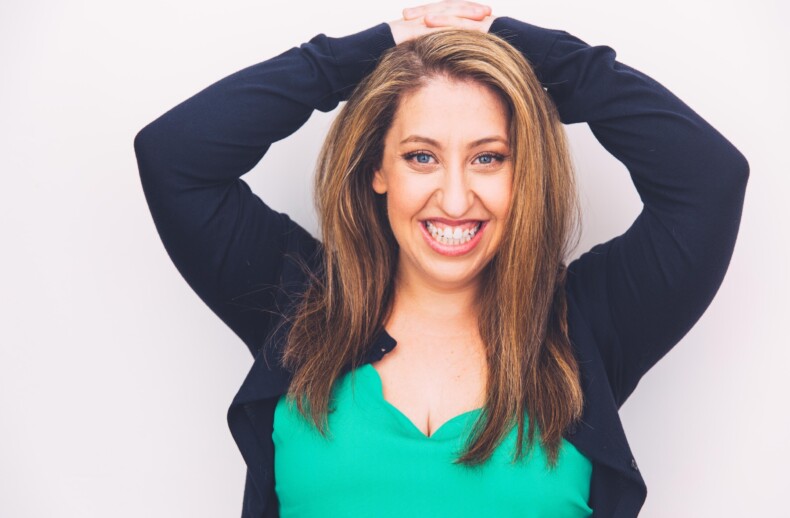 A woman with long brown hair smiles brightly against a white background. She wears a green top and black cardigan, her hands resting on her head. Her demeanor radiates warmth, much like an engaging facilitator of social emotional learning activities or diversity programs.