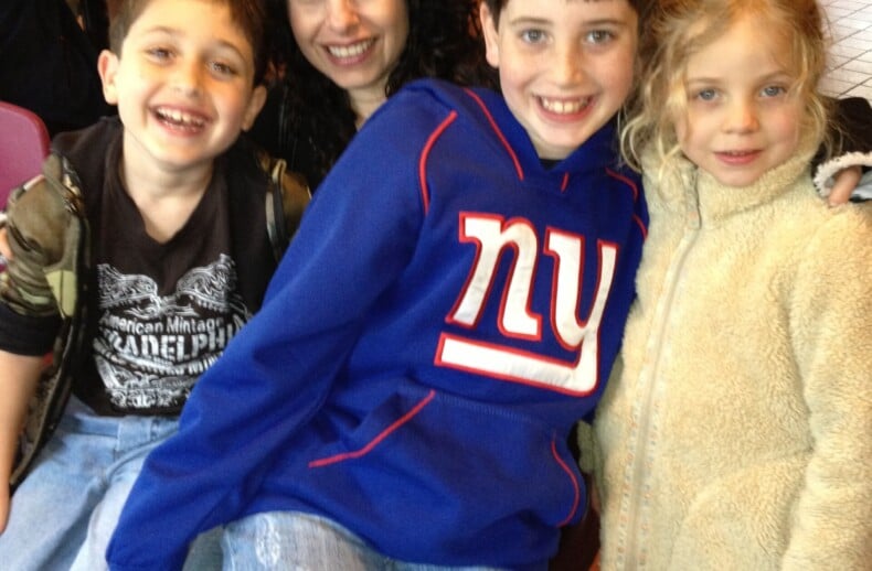 A woman with long dark hair sits in a restaurant, her lap filled with three smiling children. The boy on the left wears a dark t-shirt, the one in the middle sports a blue hoodie, and the girl on the right is cozy in her beige fleece jacket, embodying the warmth of SEL activities.