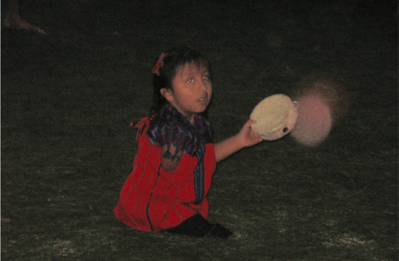 A child in a colorful outfit kneels on the grass, holding a round object in one hand. It is nighttime, and the scene is dimly lit. The child's expression of curiosity mirrors themes from an SEL curriculum, as they gaze upward with wonder.