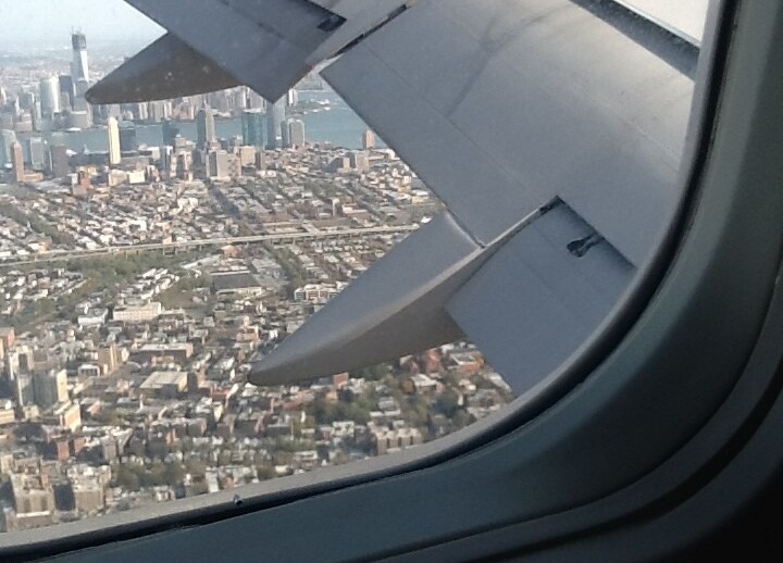 View from an airplane window showing a vibrant cityscape below, with tall buildings and a prominent skyscraper near a body of water—a scene that sparks curiosity akin to engaging in get-to-know-you activities for students. The airplane's wing is visible in the foreground, framing the city's diversity.