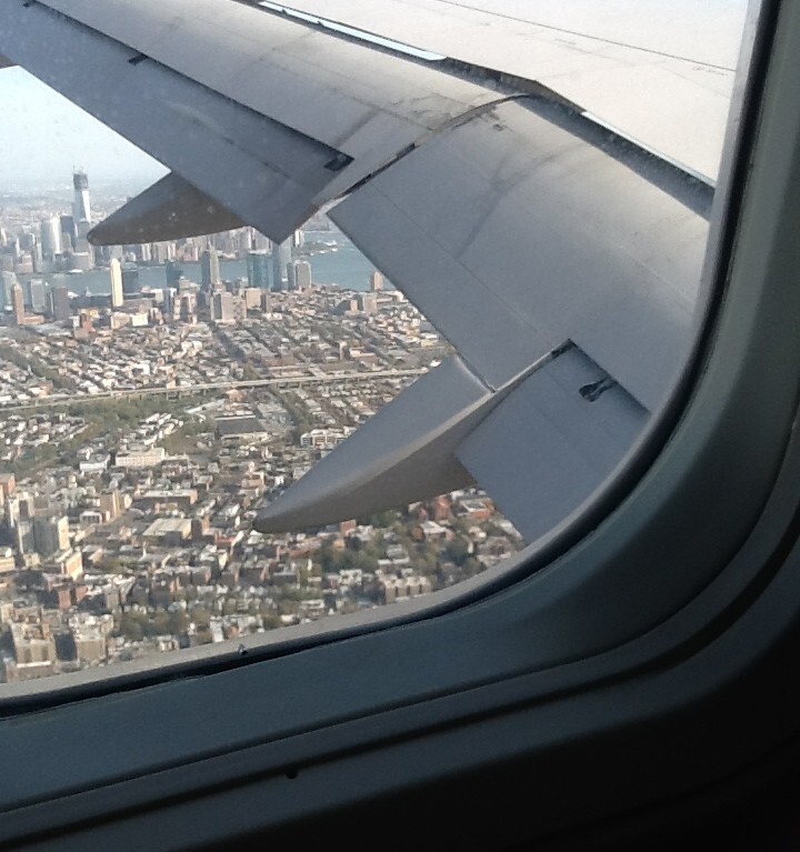 View from an airplane window showing a vibrant cityscape below, with tall buildings and a prominent skyscraper near a body of water—a scene that sparks curiosity akin to engaging in get-to-know-you activities for students. The airplane's wing is visible in the foreground, framing the city's diversity.