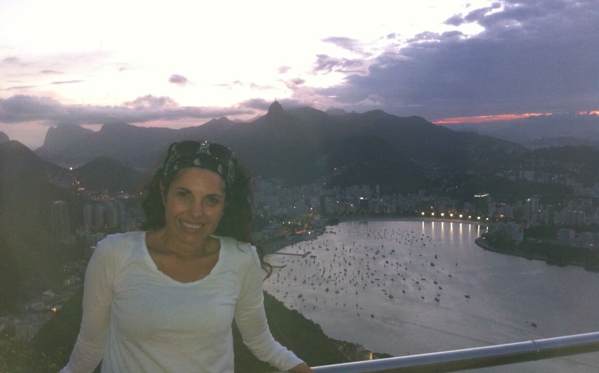 A woman in a white shirt stands smiling in front of a railing overlooking a bay dotted with boats. Behind her, the cityscape features mountains under a cloudy sky at dusk, offering inspiration akin to SEL activities that encourage emotional connection and self-awareness.