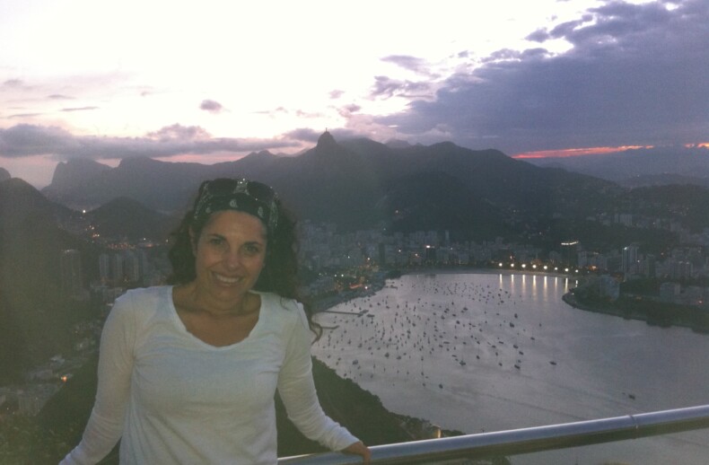 A woman in a white shirt stands smiling in front of a railing overlooking a bay dotted with boats. Behind her, the cityscape features mountains under a cloudy sky at dusk, offering inspiration akin to SEL activities that encourage emotional connection and self-awareness.