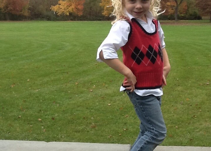 A young girl with curly blonde hair poses confidently on a stone ledge in a park, embodying the spirit of social emotional learning activities. She wears a red argyle vest over a white shirt, jeans, and black shoes. In the background, trees display colorful autumn foliage.