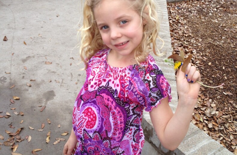A smiling young girl with curly blond hair enjoys a play moment outdoors, incorporating SEL activities into her day. She's wearing a colorful pink and purple dress, holding a yellow flower and a leaf amid scattered leaves. A green trash can is subtly present in the background.