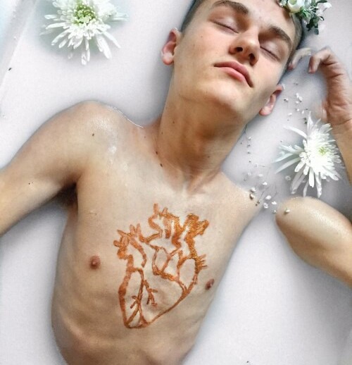 A person is lying in a milky bath with eyes closed, wearing a floral crown. White flowers float around, and a stylized heart design is painted on the chest—a moment of reflection akin to SEL activities that nurture inner peace.