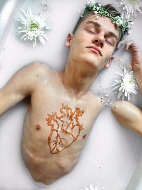 A person is lying in a milky bath with eyes closed, wearing a floral crown. White flowers float around, and a stylized heart design is painted on the chest—a moment of reflection akin to SEL activities that nurture inner peace.