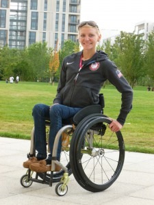 A woman in a wheelchair smiles outdoors, embodying the spirit of diversity programs. She's wearing a dark jacket with a U.S. emblem, jeans, and sunglasses perched on her head. The background showcases green grass, trees, and modern buildings.