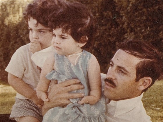 A man with a mustache is holding two young children, capturing the spirit of social emotional learning activities. The girl in a blue dress and the boy in a light-colored shirt and shorts stand together outdoors, amidst lush greenery that enhances their connection.