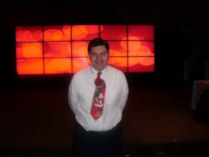 A person wearing a white shirt and a festive tie stands in front of a large digital display showing red and orange abstract patterns, where social emotional learning activities are creatively integrated for students.