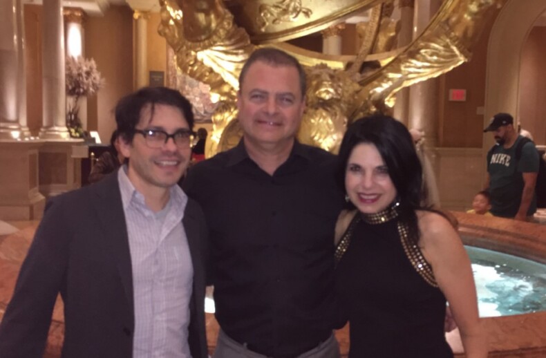 Three people pose for a picture in an elegant setting, featuring a decorative sculpture in the background. As part of diversity programs, they stand before a marble fountain with golden accents, smiling and dressed in semi-formal attire, celebrating unity and inclusion.