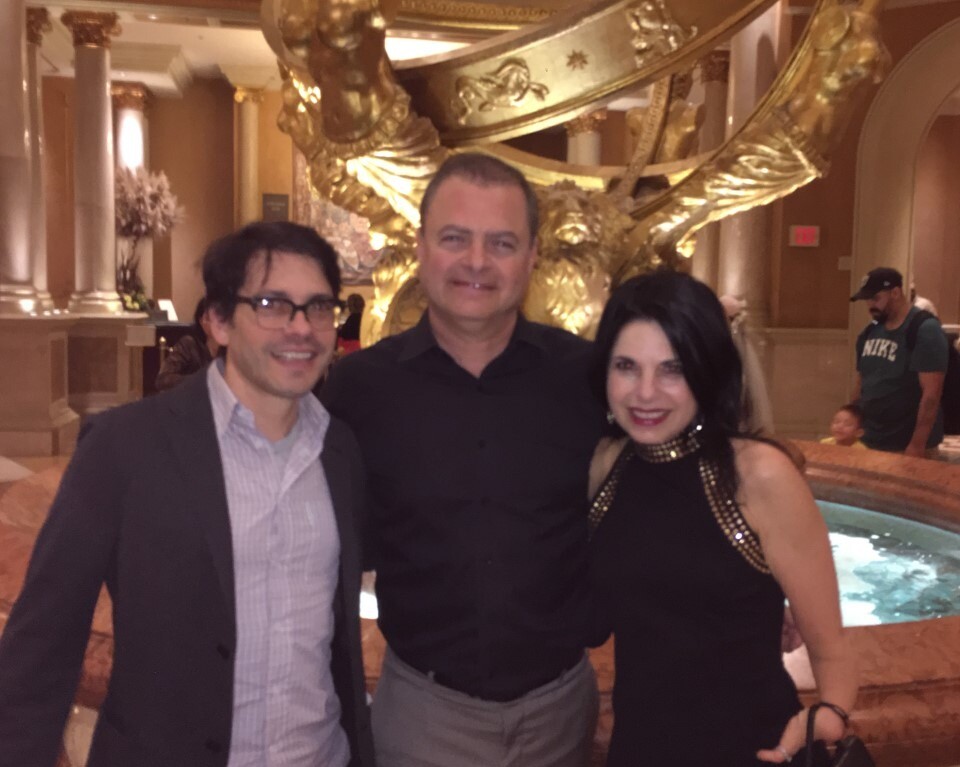 Three people pose for a picture in an elegant setting, featuring a decorative sculpture in the background. As part of diversity programs, they stand before a marble fountain with golden accents, smiling and dressed in semi-formal attire, celebrating unity and inclusion.