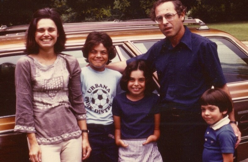 A family of five stands in front of a brown station wagon, embodying the essence of SEL activities. The woman wears a patterned blouse, and the man sports glasses and a dark shirt. The two older children stand in the middle, while the youngest is next to the man. Lush trees grace the background.