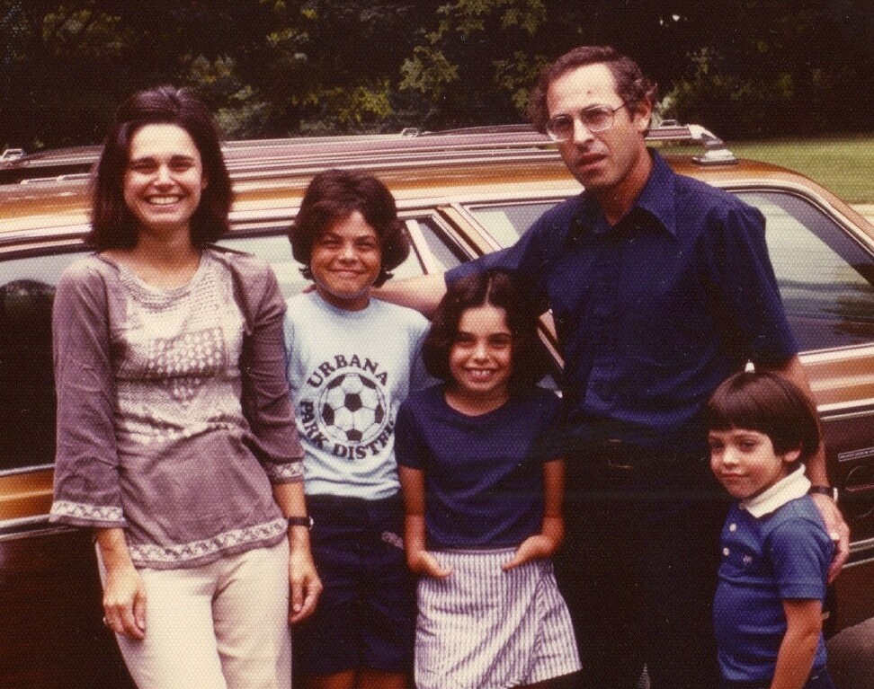 A family of five stands in front of a brown station wagon, embodying the essence of SEL activities. The woman wears a patterned blouse, and the man sports glasses and a dark shirt. The two older children stand in the middle, while the youngest is next to the man. Lush trees grace the background.