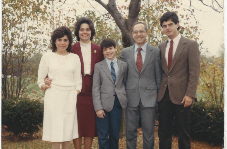 A family of six, including four adults and two children, stands together outdoors in front of a tree. Dressed in formal attire with suits, ties, and dresses, they embody the connection emphasized by social emotional learning activities. Leaves and trees frame their gathering beautifully.