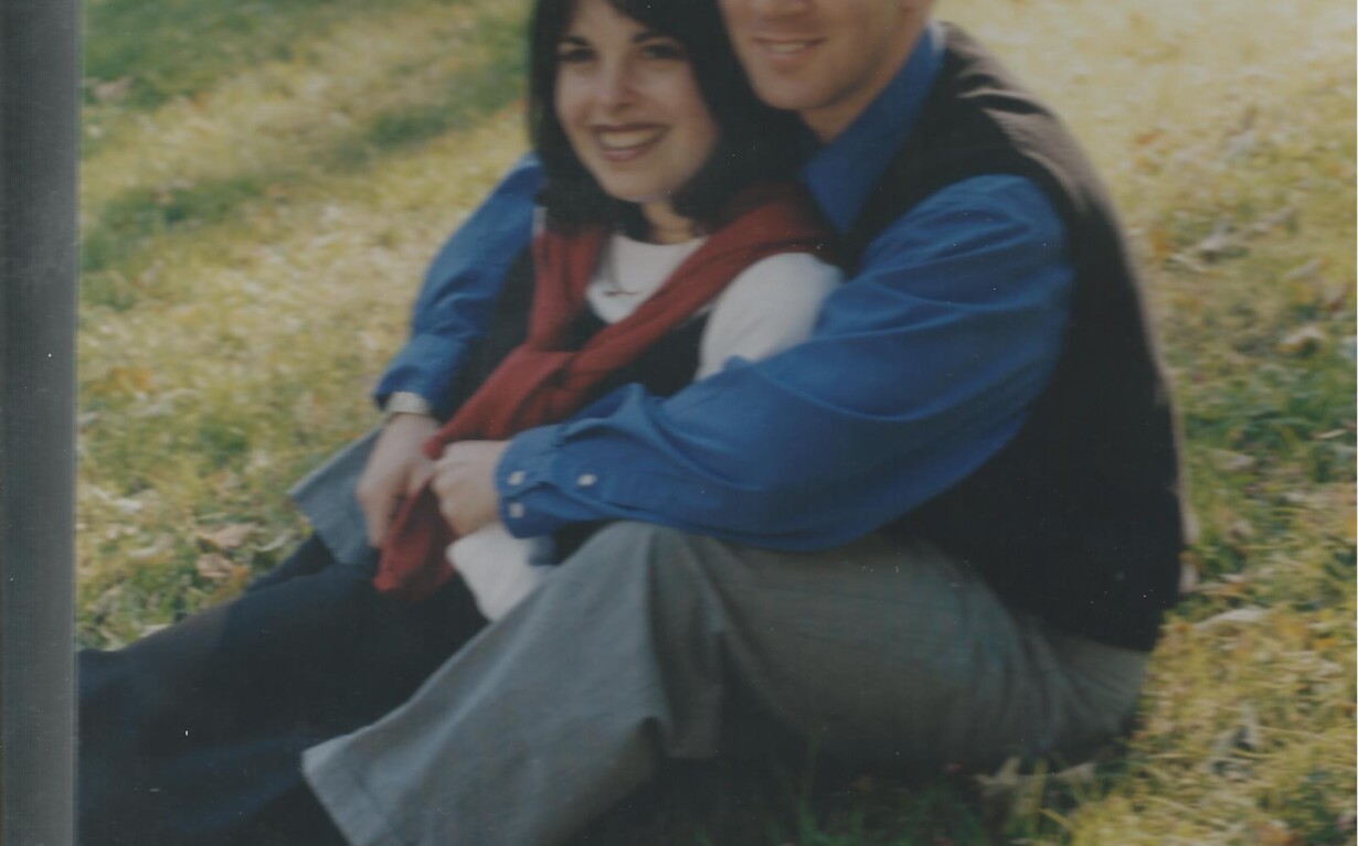 A couple sits on grass, leaning against each other and smiling, embodying the warmth of social emotional learning activities. The woman has dark hair and wears a red scarf, white top, and black pants. The man has short hair and wears a blue shirt with a black vest and gray pants. Sunlight filters through trees.