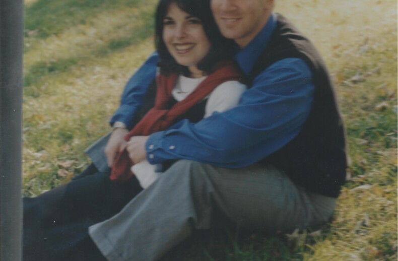 A couple sits on grass, leaning against each other and smiling, embodying the warmth of social emotional learning activities. The woman has dark hair and wears a red scarf, white top, and black pants. The man has short hair and wears a blue shirt with a black vest and gray pants. Sunlight filters through trees.