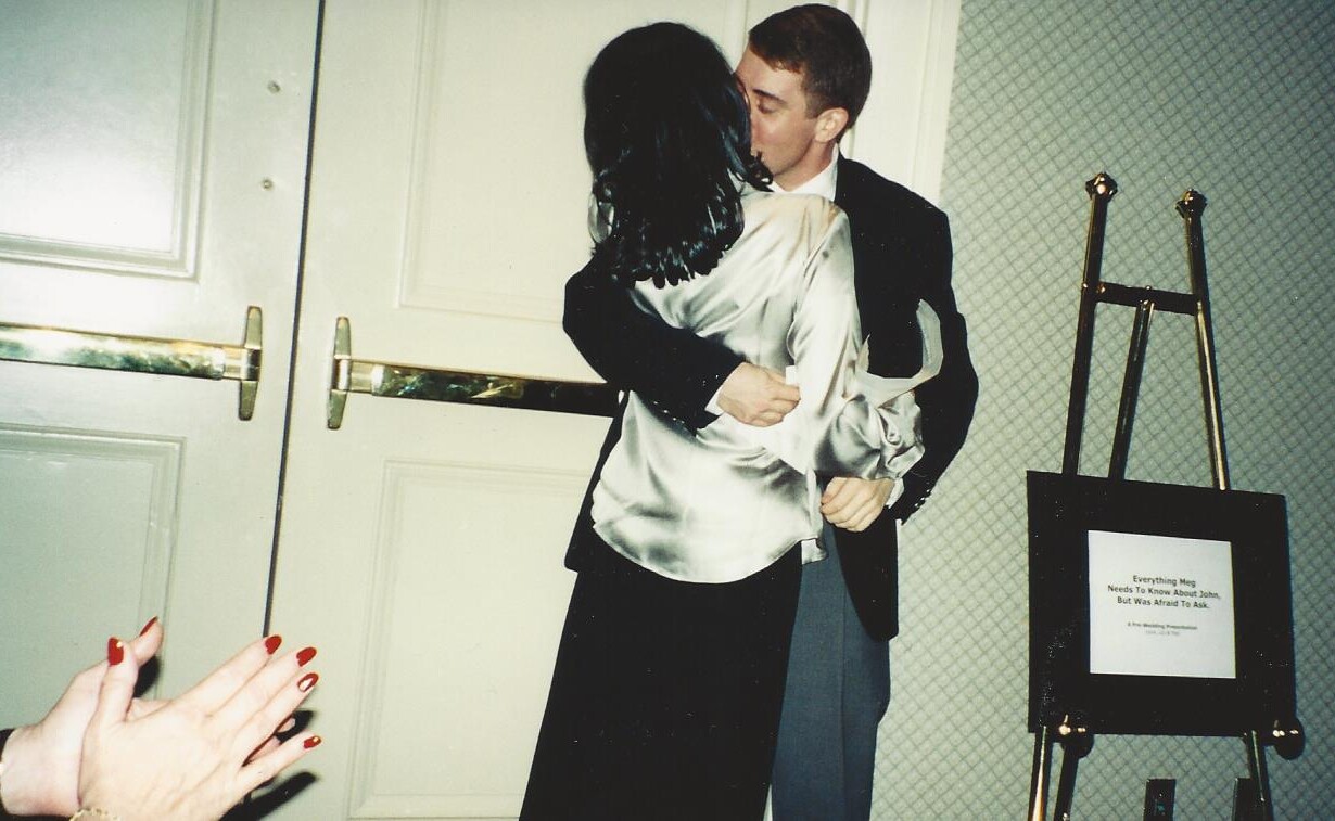 A couple shares a tender kiss in front of closed doors, with the woman in a shiny blouse and the man in a suit. A framed sign rests on an easel to their right as hands with red-painted nails clap nearby, capturing an unexpected moment of connection fit for any SEL curriculum's focus on emotions.