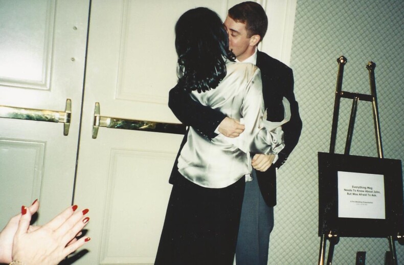 A couple shares a tender kiss in front of closed doors, with the woman in a shiny blouse and the man in a suit. A framed sign rests on an easel to their right as hands with red-painted nails clap nearby, capturing an unexpected moment of connection fit for any SEL curriculum's focus on emotions.