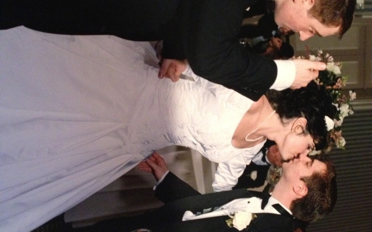 A bride and groom in a wedding dress and tuxedo share a kiss beneath a floral arch indoors. Beside them, a man in a dark suit, resembling an instructor of social emotional learning activities, holds a microphone and champagne flute. Enthusiastic guests are visible in the background.