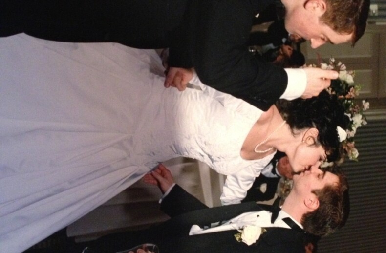 A bride and groom in a wedding dress and tuxedo share a kiss beneath a floral arch indoors. Beside them, a man in a dark suit, resembling an instructor of social emotional learning activities, holds a microphone and champagne flute. Enthusiastic guests are visible in the background.