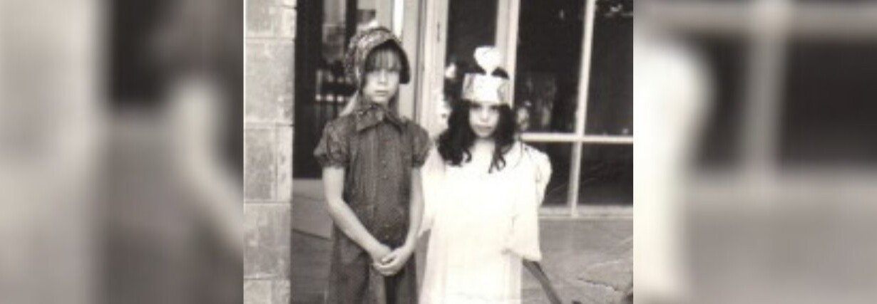 Two children in costume, engaging in SEL activities, stand outside a building. One wears a dark outfit with a patterned hat, while the other sports a light dress and paper crown. A large window and part of a stone wall form the backdrop of this black-and-white image.