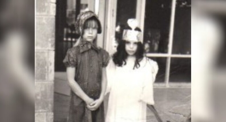 Two children in costume, engaging in SEL activities, stand outside a building. One wears a dark outfit with a patterned hat, while the other sports a light dress and paper crown. A large window and part of a stone wall form the backdrop of this black-and-white image.
