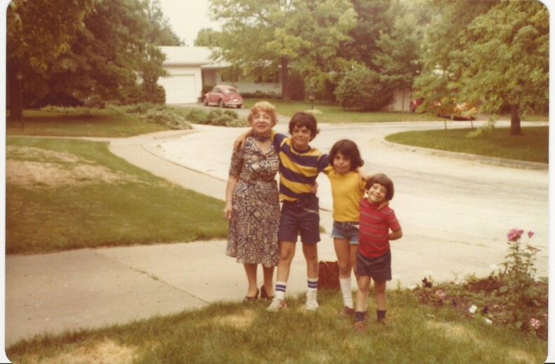 A woman stands smiling with three children on a suburban street, embodying the values of social emotional learning. The children, dressed casually in colorful attire, playfully engage with their surroundings. Behind them, trees and a car add to this lively scene of connection and growth.