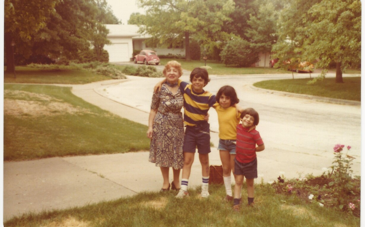 A woman stands arm in arm with three children on a suburban street, embodying the essence of diversity programs. She wears a patterned dress, and the children are in casual summer clothing. A grassy lawn and trees frame the scene, alongside houses and parked cars.