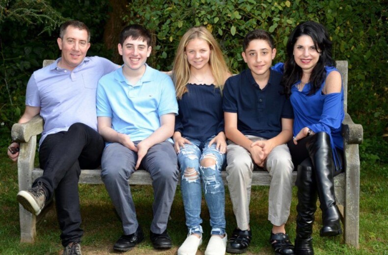 A family of five sits on a wooden bench outdoors, embodying the warmth found in social emotional learning. Two adults are on the ends with three children between them, all smiling in blue and black tones. Green foliage flourishes in the background, highlighting their connection to nature and each other.