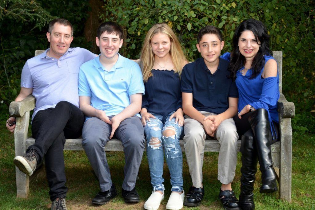 A family of five sits on a wooden bench outdoors, embodying the warmth found in social emotional learning. Two adults are on the ends with three children between them, all smiling in blue and black tones. Green foliage flourishes in the background, highlighting their connection to nature and each other.