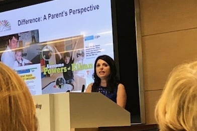 A woman is speaking at a podium during a presentation titled "Difference: A Parent's Perspective." The slide behind her shows family photos and text, including "Awesome Powers - Aidan." Audience members are partially visible in the foreground.