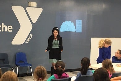 A woman stands in front of a group of seated children in a room with a blue wall displaying the YMCA logo. She appears to be speaking or presenting. Two children sit at a table on the right, engaged in an activity.