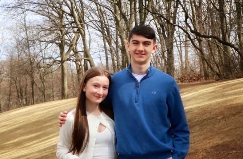 A young woman and man standing outside, smiling at the camera. They are wearing casual clothing; she is in a white top and he is in a blue sweater. Bare trees and a grassy area fill the background.