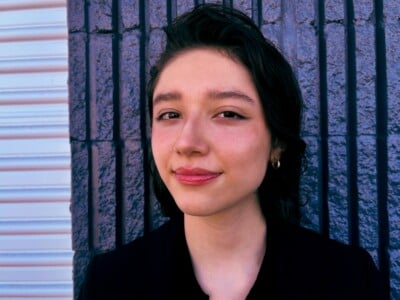 A young person with short dark hair and a slight smile stands in front of a textured wall with dark vertical tiles and a white section on the left, wearing a black top and small hoop earrings.