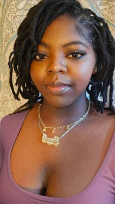 A woman with long, styled dreadlocks wearing a mauve top and layered gold necklaces stands in front of a patterned wallpaper, looking at the camera with a slight smile.