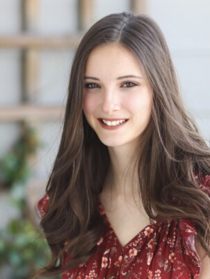 A young woman with long brown hair and fair skin smiles at the camera. She is wearing a red floral top and is standing in front of a blurred background with greenery and wooden lattice.
