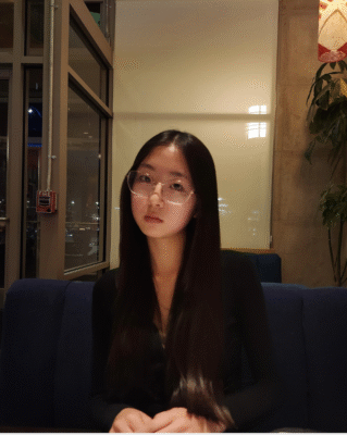 A woman with long, straight dark hair and glasses sits at a blue booth in a dimly lit restaurant, facing the camera with a neutral expression. A plant and lamp are visible in the background.